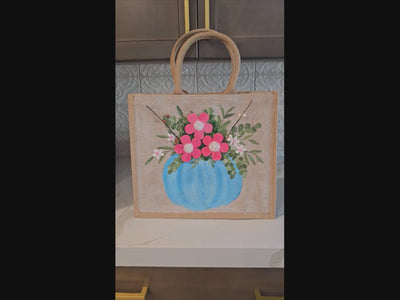 Hand-painted jute tote featuring a blue pumpkin used as a flower pot, filled with pink flowers and greenery; painted on off-white metallic background with beads, leaves, and wooden floral embellishments