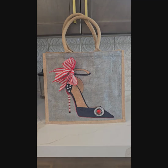 Video of hand-painted stiletto design featuring white stars, a red-and-white striped heel, a striped bow, and a sparkling red jewel embellishment on a silver tote bag.