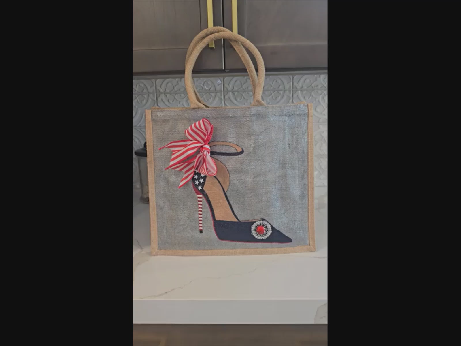 Video of hand-painted stiletto design featuring white stars, a red-and-white striped heel, a striped bow, and a sparkling red jewel embellishment on a silver tote bag.