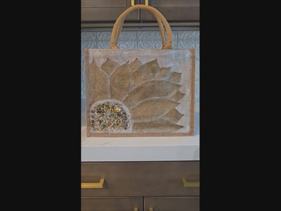 Video of Hand-painted neutral sunflower tote bag featuring soft beige and taupe petals with pearl and gold jeweled embellishments at the flower center, accented with natural jute trim and rounded padded handles against a neutral background.