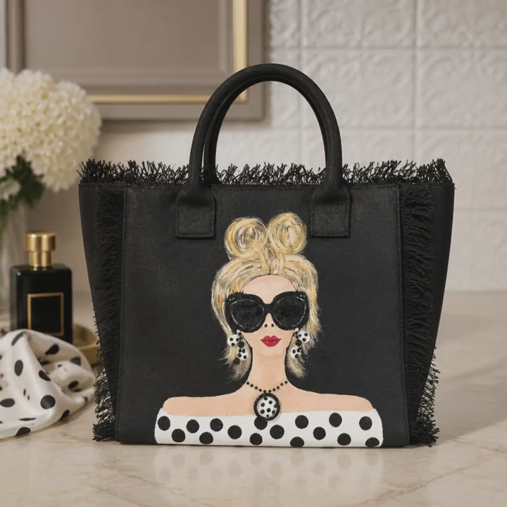 video of Hand-painted black fringed canvas tote bag featuring a blonde woman wearing oversized sunglasses, polka dot earrings, and a matching polka dot top with dimensional jewelry embellishments.