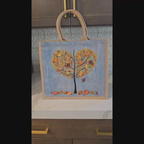 Hand-painted jute tote  featuring a heart-shaped fall tree with colorful leaves in red, orange, and gold; painted on a pale blue metallic background with beads and wooden embellishments