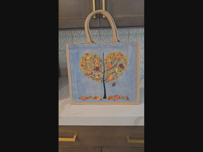 Hand-painted jute tote  featuring a heart-shaped fall tree with colorful leaves in red, orange, and gold; painted on a pale blue metallic background with beads and wooden embellishments
