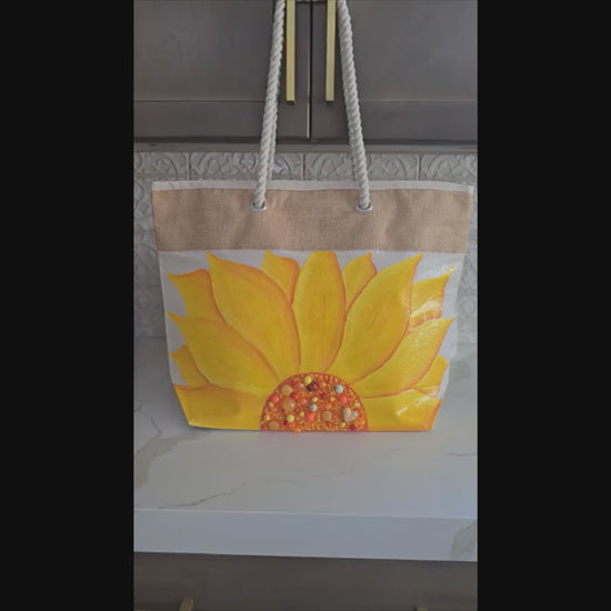 Video of Hand-painted rope handle beach tote featuring a large dimensional yellow sunflower design with textured petals and embellished center details on a natural jute and canvas bag with cream rope handles.