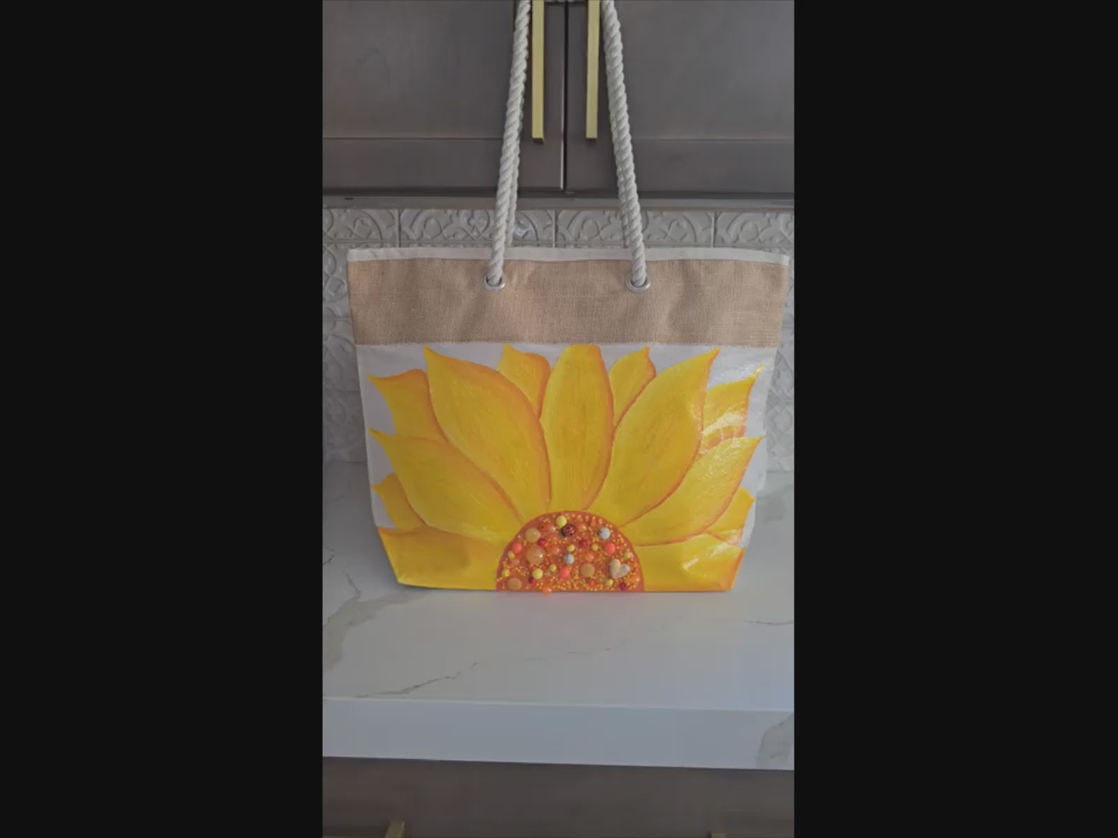 Video of Hand-painted rope handle beach tote featuring a large dimensional yellow sunflower design with textured petals and embellished center details on a natural jute and canvas bag with cream rope handles.