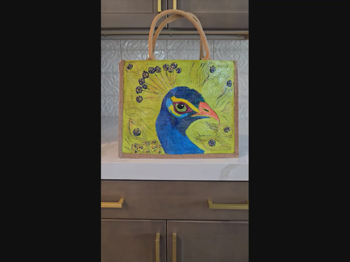 Video ofHand-painted peacock tote bag on natural jute canvas featuring a bold blue peacock portrait with colorful feather accents against a vibrant lime green background, finished with long double handles and a structured rectangular shape. 