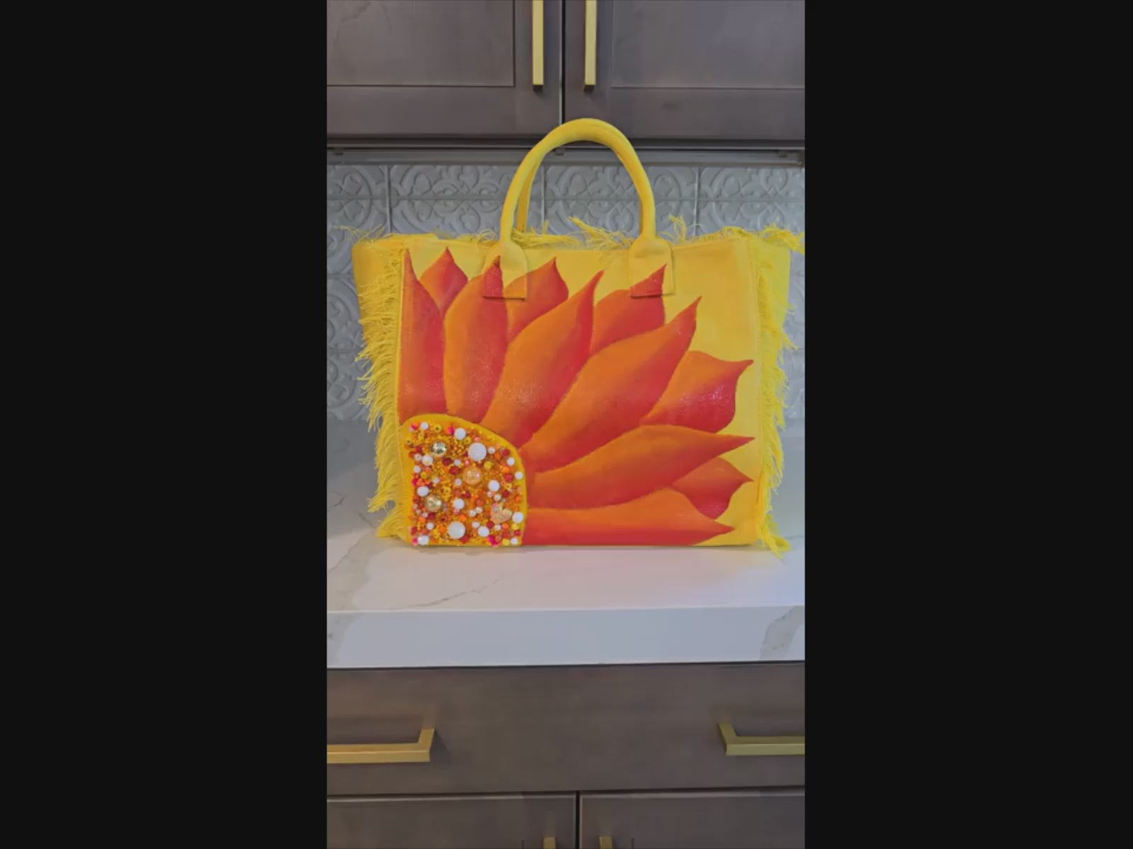 video of Hand-painted yellow fringed tote bag featuring a bold sunflower design in vibrant orange and red tones, accented with pearl and gold embellishments on the flower center, displayed against a neutral background.