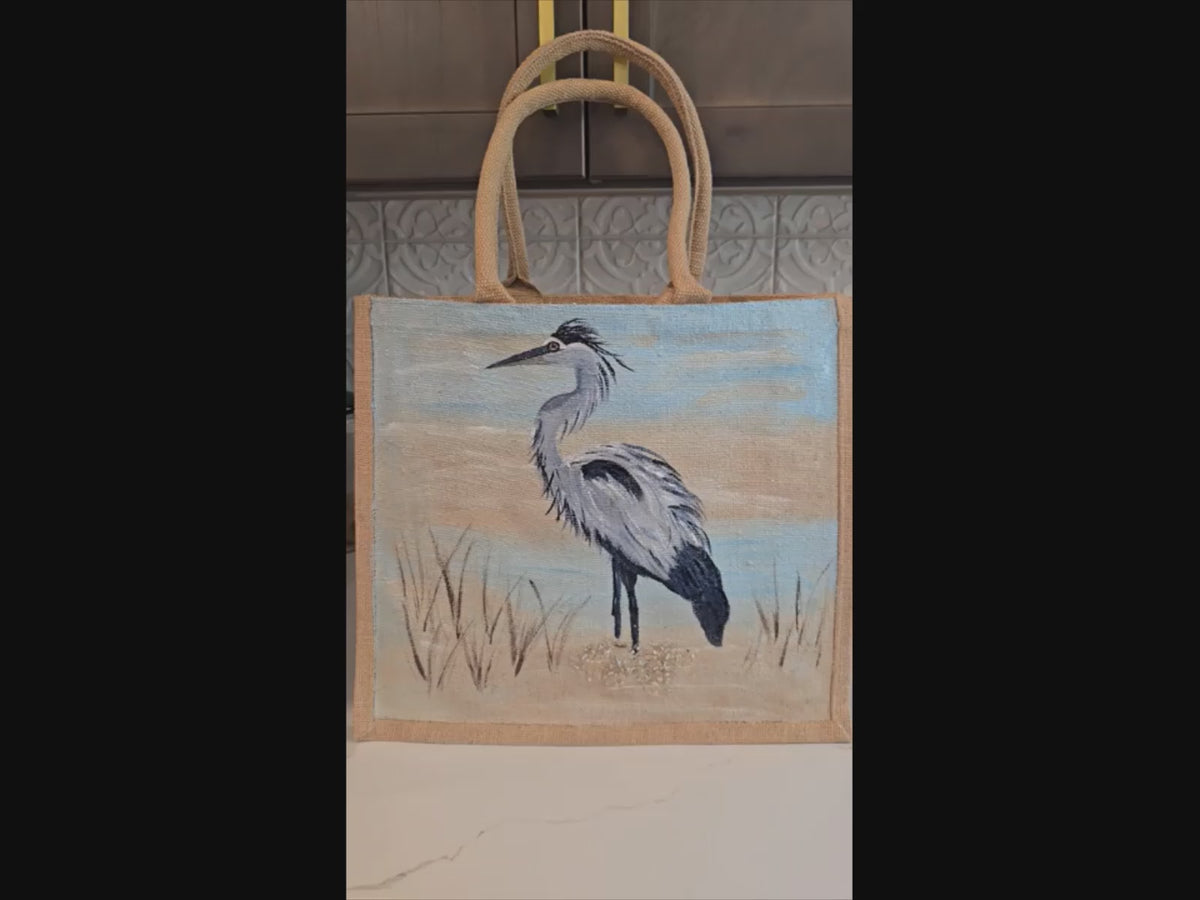 Video of hand painted blue heron with a beachy background embellished with glitter glass on jute tote