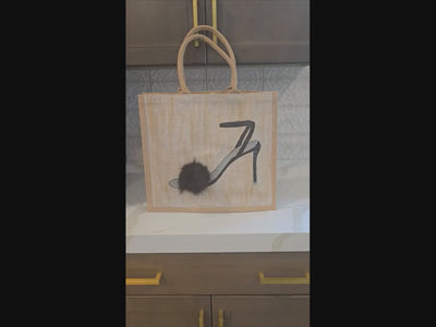 video of Hand-painted jute tote bag featuring a black strappy high heel sandal with a soft black fur pom accent on a neutral textured background. The tote has natural woven handles and a chic, fashion-inspired design with minimalist black and beige tones.