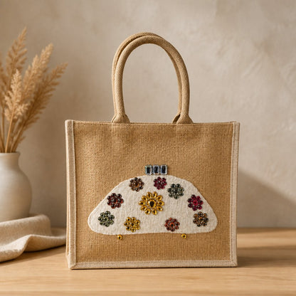 Hand-painted natural jute tote bag featuring a white half-moon purse design embellished with colorful jewel-like floral accents in gold, red, green, and amber tones. The structured tote has rounded woven handles and a neutral background, giving it an elegant artisan-inspired look.