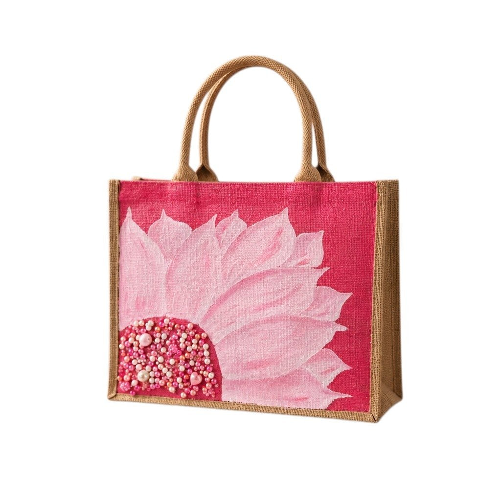 Hand-painted jute tote bag featuring a large soft pink flower with layered petals and a jeweled center, accented with pearl and bead embellishments on a vibrant pink background.