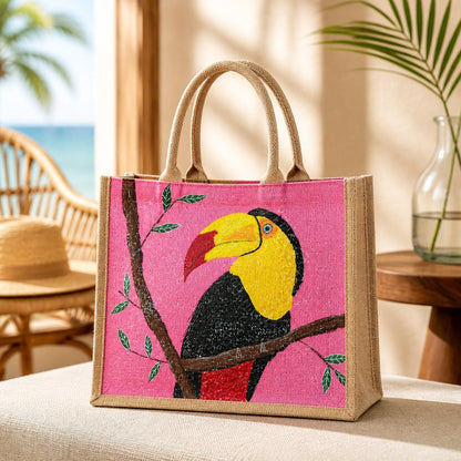 Hand-painted jute tote bag featuring a colorful toucan perched on a branch against a bright pink background, with natural burlap sides and handles.