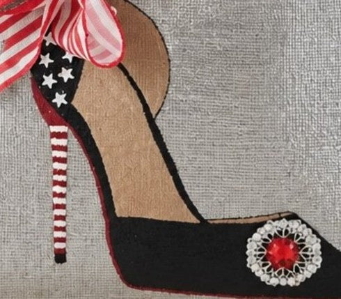 Close-up of a hand-painted stiletto design featuring white stars, a red-and-white striped heel, a striped bow, and a sparkling red jewel embellishment on a silver tote bag.